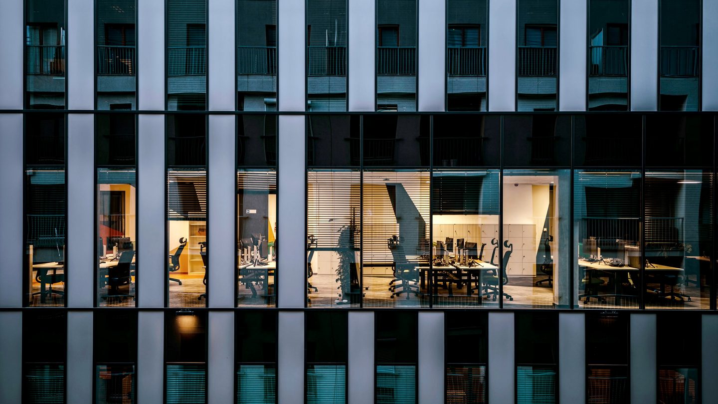 An office building at dusk with lit workspaces visible through the glass